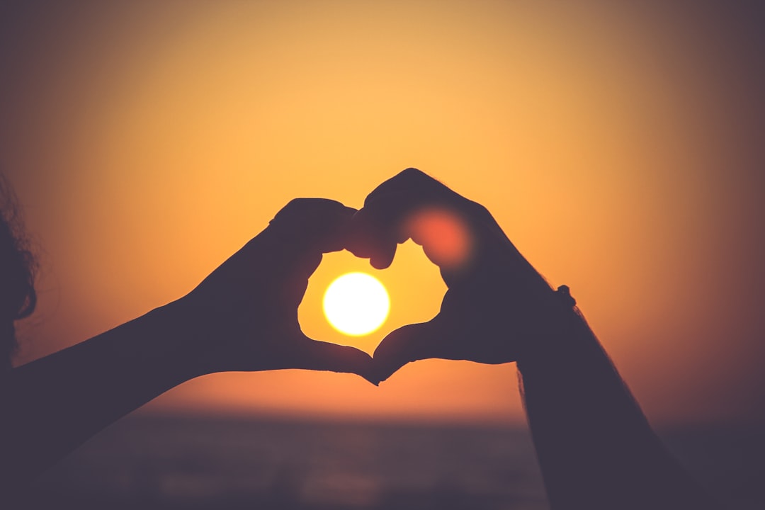 Two hands making the shape of a heart in front of a sunset.